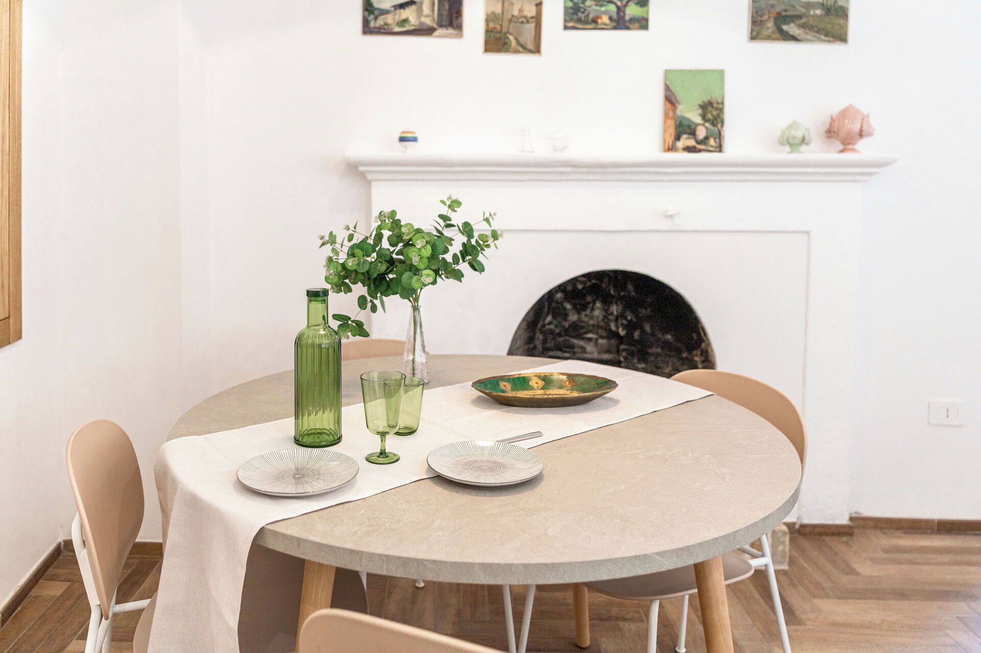 A dining table with a vase of green plants, two glasses, and a plate with a green bowl.