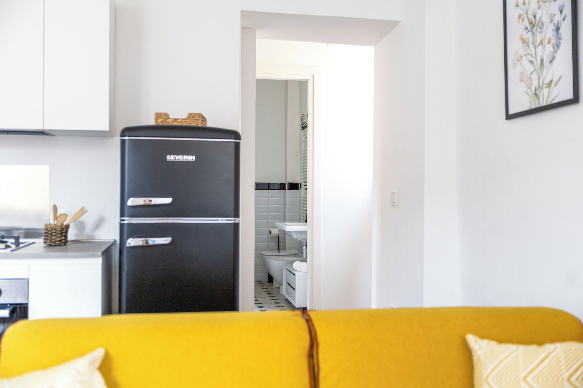 A compact kitchen with a black refrigerator, a sink, and a toilet visible through an open doorway.