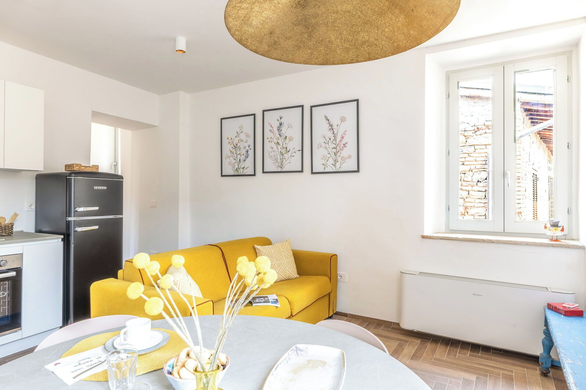 Ein modernes Wohnzimmer mit einem gelben Sofa, einem Esstisch mit einer Vase voller gelber Blumen und einer Küchenzeile mit einem schwarzen Kühlschrank.