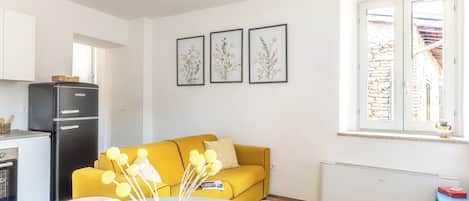 A modern living room with a yellow sofa, a dining table with a vase of yellow flowers, and a kitchen area with a black refrigerator.