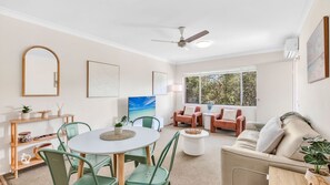 Un salon bien éclairé, avec une table ronde, des chaises vertes, un canapé beige et un ventilateur de plafond.
