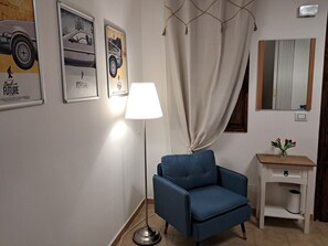 A room with a blue armchair, a floor lamp, a small table with a vase, and two posters on the wall.