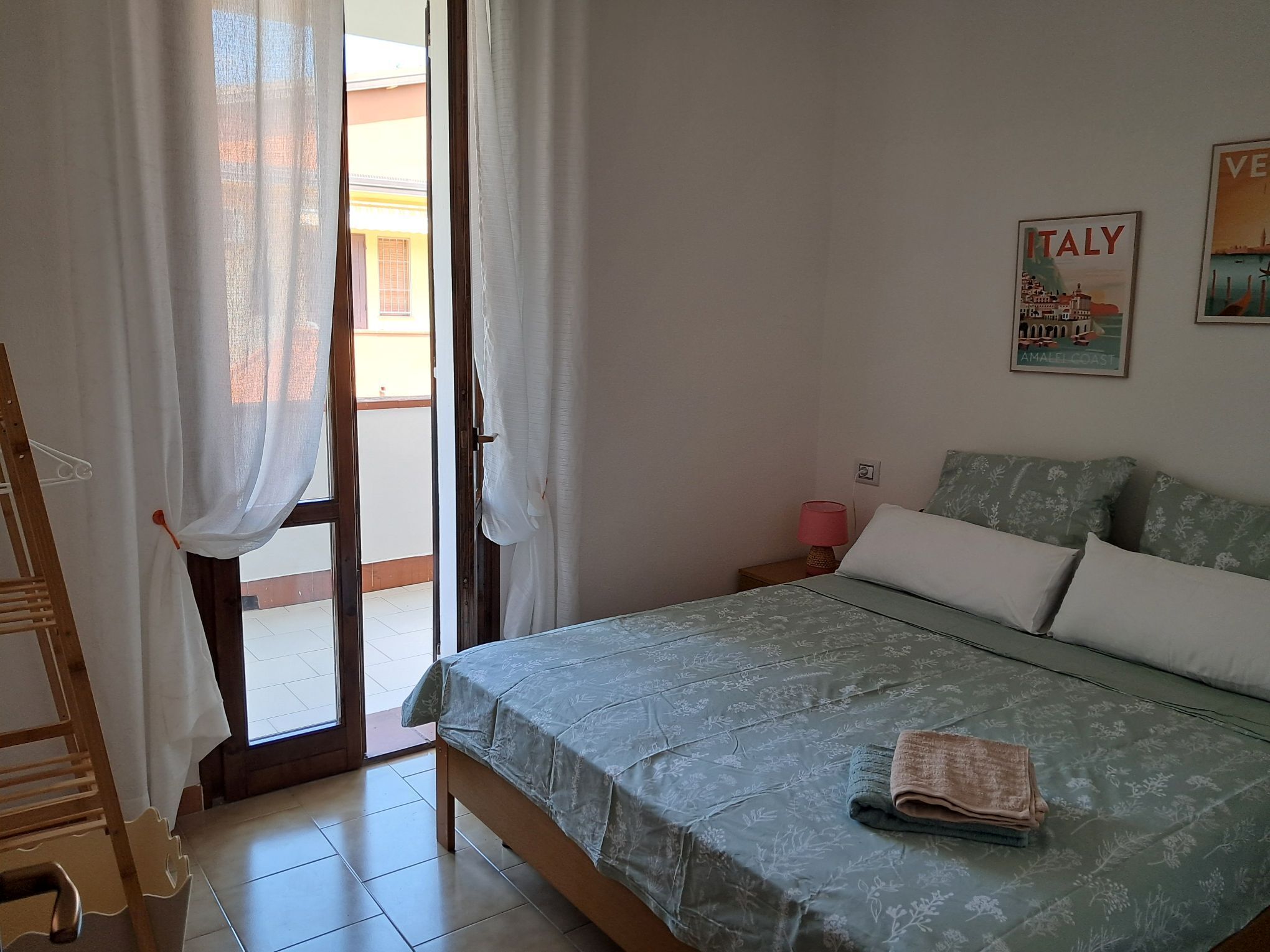 A bedroom with a bed, a nightstand, a lamp, a poster on the wall, and a view of the outside.