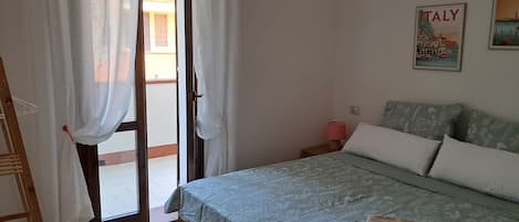 A bedroom with a bed, a nightstand, a lamp, a poster on the wall, and a view of the outside.
