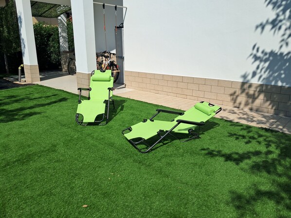 Two green lawn chairs on artificial grass, a white pillar, and a brick wall.
