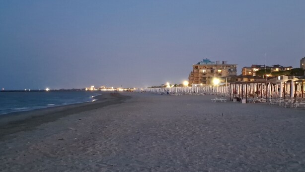 A beach with a row of beach umbrellas and chairs, a building in the background, and a calm sea.