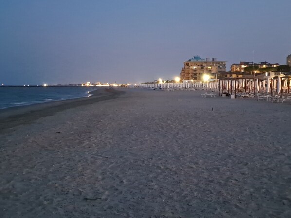 A beach with a row of beach umbrellas and chairs, a building in the background, and a calm sea.