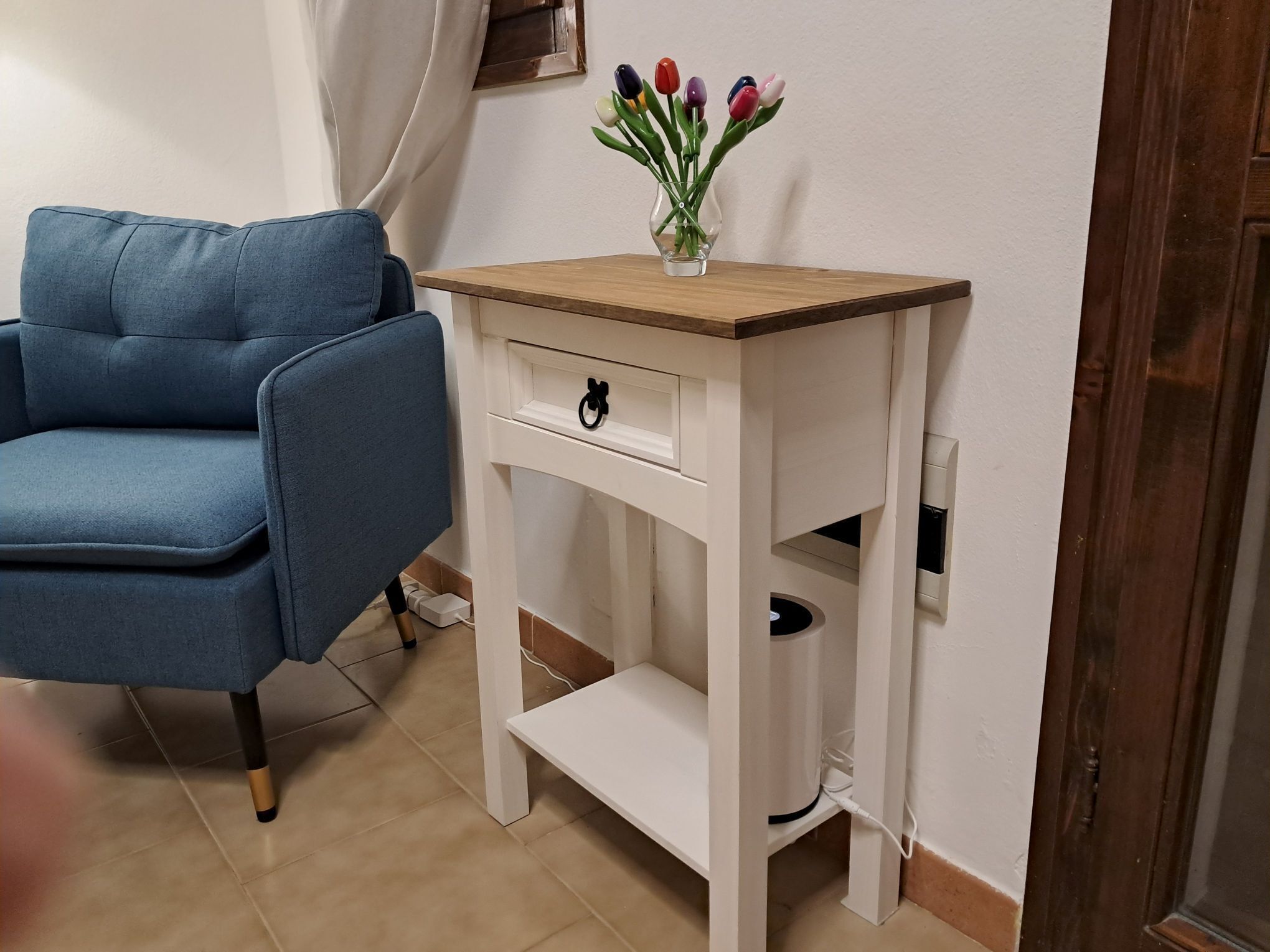 A room with a blue armchair, a white side table with a vase of flowers, and a wooden door.
