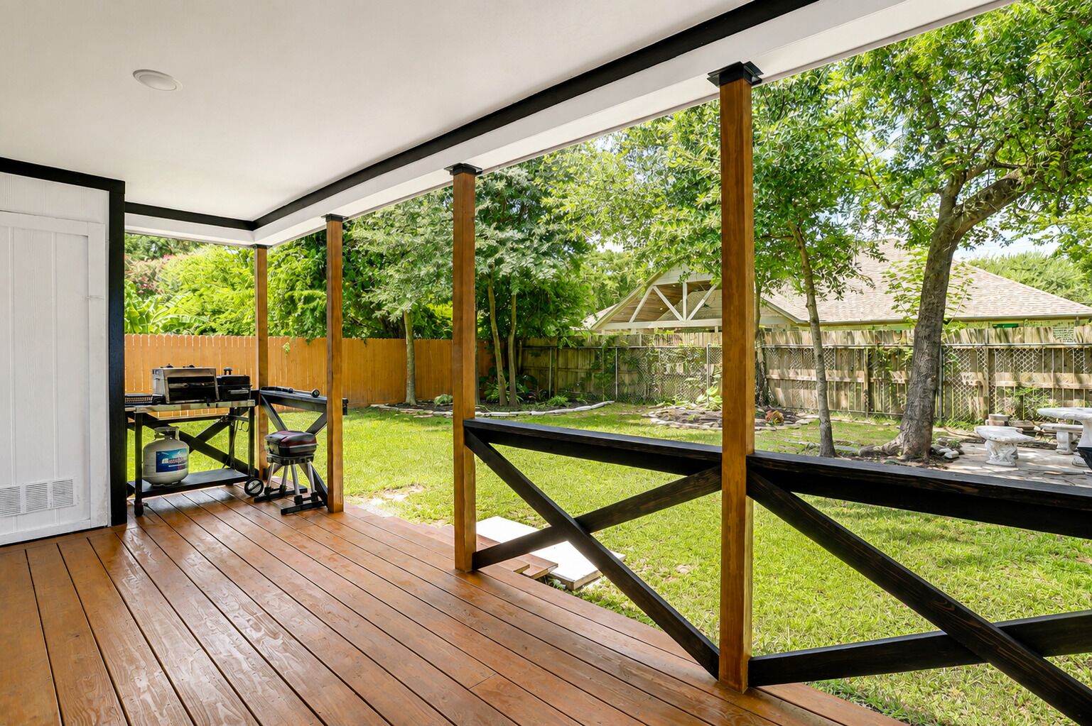 A covered patio with a wooden deck, a grill, and a view of a backyard with a wooden fence and trees.