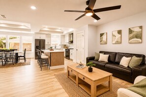 A modern kitchen with a central island, a dining area with a wooden table and chairs, a living room with a brown leather sofa, and a ceiling fan.