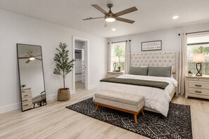 A bedroom with a bed, a ceiling fan, a mirror, a potted plant, and a bedside table with a lamp.