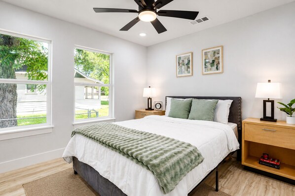 Un dormitorio con cama, mesitas de noche, ventilador de techo, dos ventanas y vistas al exterior.