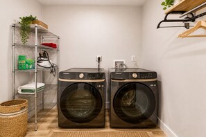A laundry room with two Samsung washing machines, a metal shelving unit with various items, and a hanging rack. - Cozy Modern Tomball Home, Family-Friendly & Spacious Backyard (Tomball)