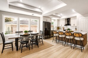A modern kitchen with a dining area, featuring a central island with bar stools, a refrigerator, and a large window providing natural light. - Cozy Modern Tomball Home, Family-Friendly & Spacious Backyard (Tomball)