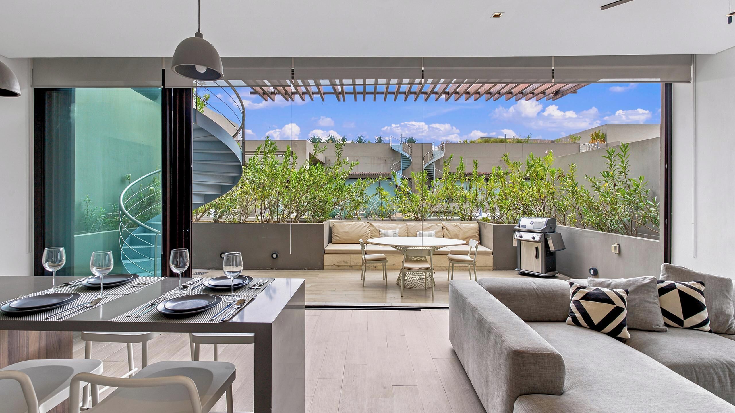 Un espace de vie moderne comprenant un coin repas, un canapé et une vue sur un escalier en colimaçon et la verdure environnante.