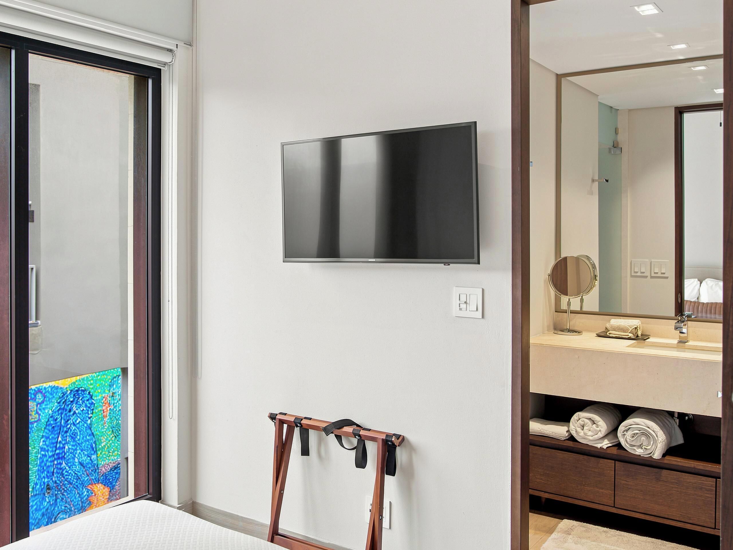 Een moderne badkamer met een flatscreen televisie aan de muur, een houten handdoekrek en een wastafelmeubel met spiegel en opbergruimte voor handdoeken.