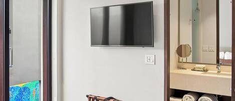 Een moderne badkamer met een flatscreen televisie aan de muur, een houten handdoekrek en een wastafelmeubel met spiegel en opbergruimte voor handdoeken.