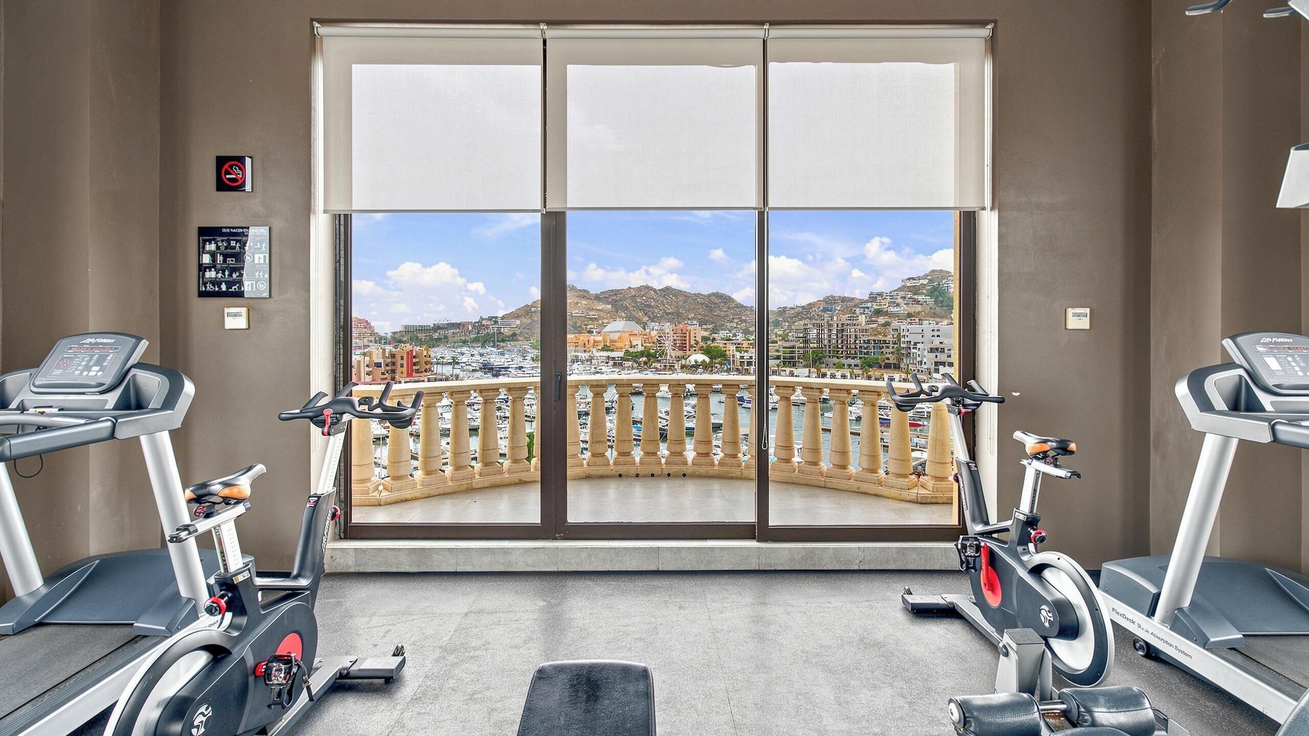 A gym with two treadmills, a large window with a view of a marina and buildings, and a sign indicating no smoking.