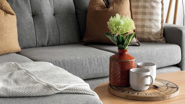 Un canapé gris avec une couverture, un vase rempli de fleurs vertes et deux tasses posées sur une table ronde en bois.