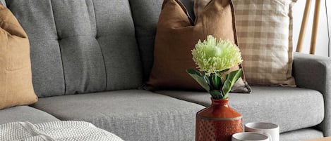 Un canapé gris avec une couverture, un vase rempli de fleurs vertes et deux tasses posées sur une table ronde en bois.