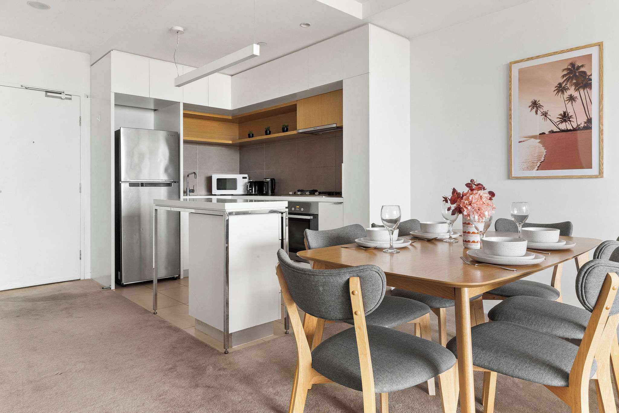 A modern kitchen with a refrigerator, microwave, and stove. There is a dining area with a wooden table and grey upholstered chairs.
