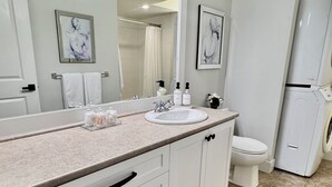 Un baño con lavamanos blanco, espejo y retrete. Hay toallas en la pared y una cortina de ducha.
