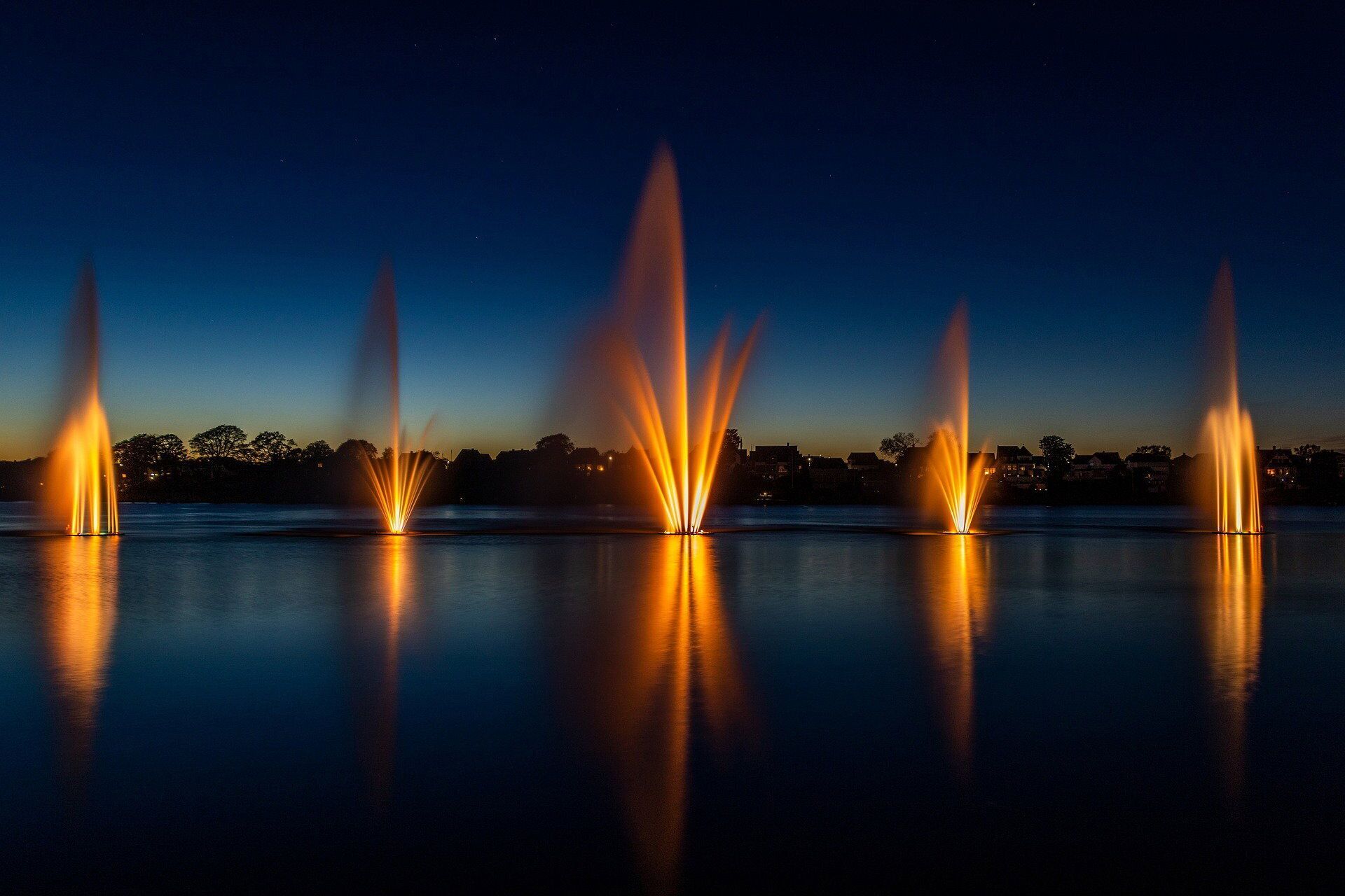 Una vista notturna di una fontana con getti d'acqua multipli, illuminata da luci che si riflettono sulla superficie.