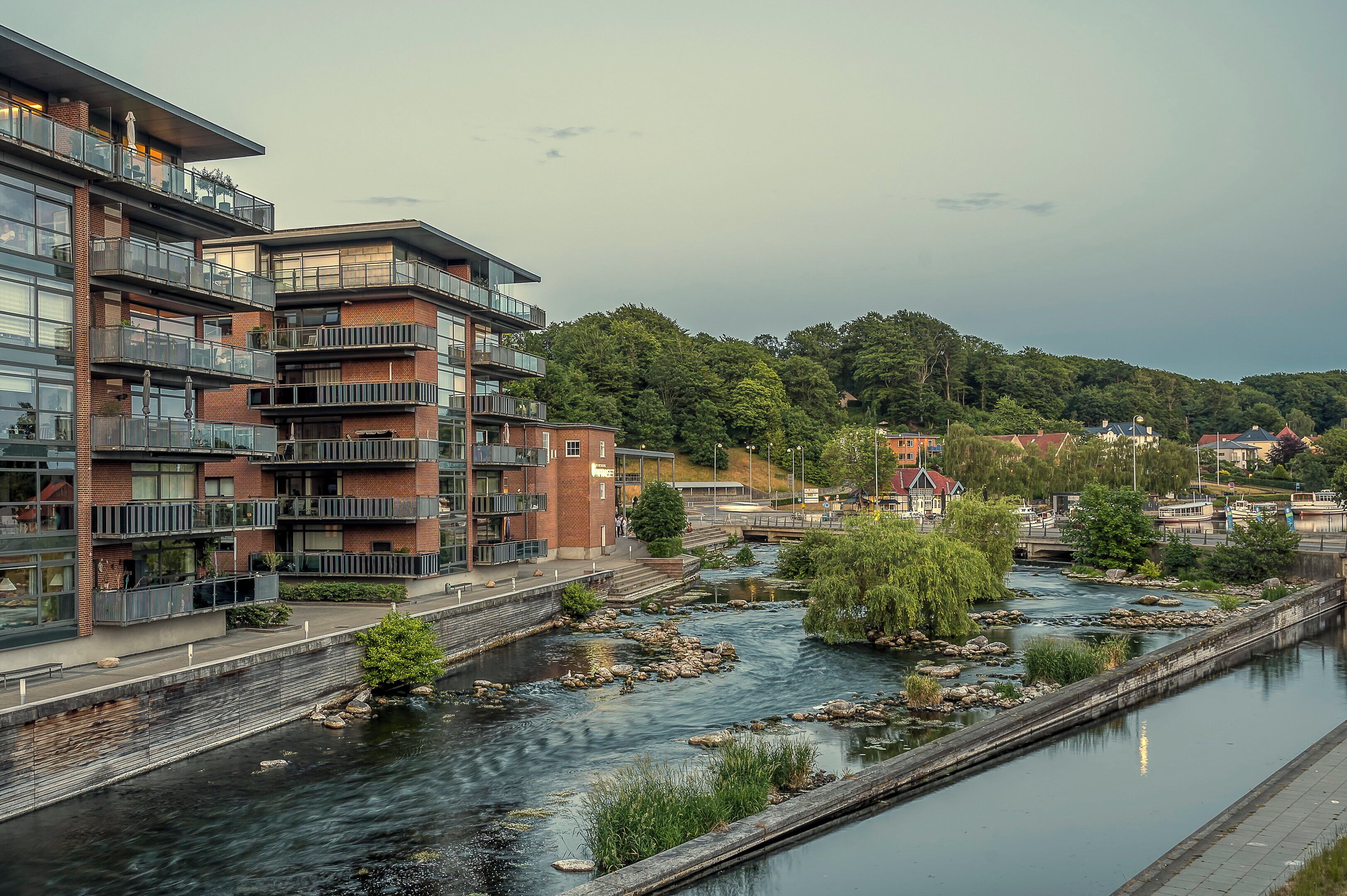 A riverside view with modern buildings, a bridge, and a forested hill.