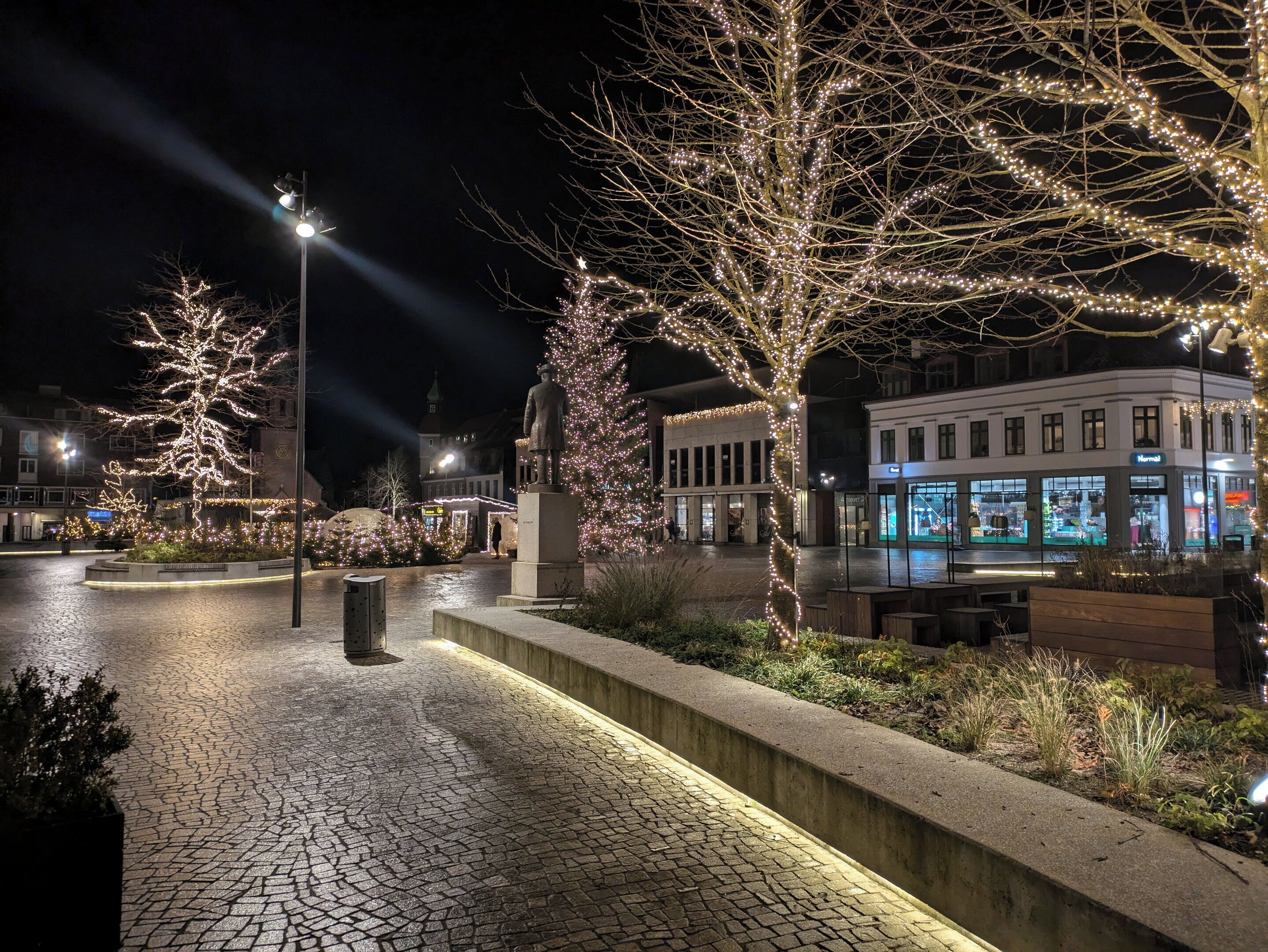 Una plaza urbana nocturna con pavimento de adoquines, árboles iluminados y una estatua.