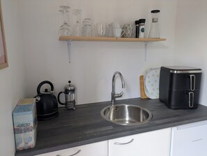 Una cocina con un lavamanos de acero inoxidable, una tetera, una cafetera, un tostador, y una repisa con cristalería y un molinillo de café.