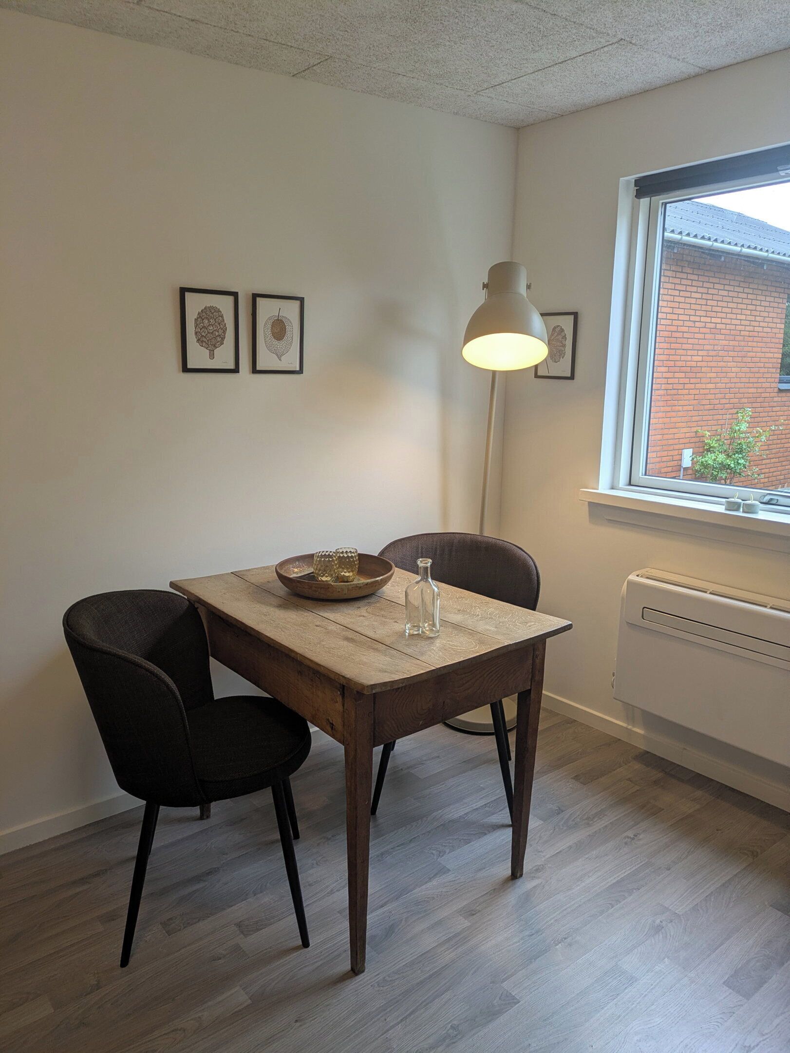 A dining area with a wooden table, two chairs, a floor lamp, and framed botanical prints on the wall.