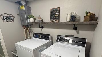 Une buanderie équipée de deux machines à laver et deux sèche-linge à chargement frontal superposés, d’un chauffe-eau et d’une étagère contenant des produits de lessive.