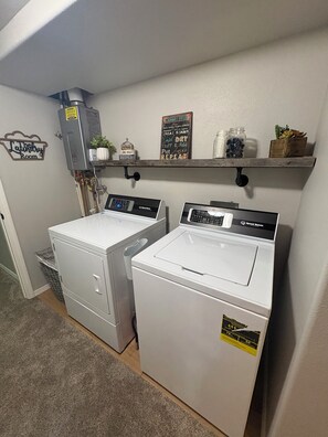 A laundry room with two stacked front-loading washers and dryers, a water heater, and a shelf with laundry supplies.