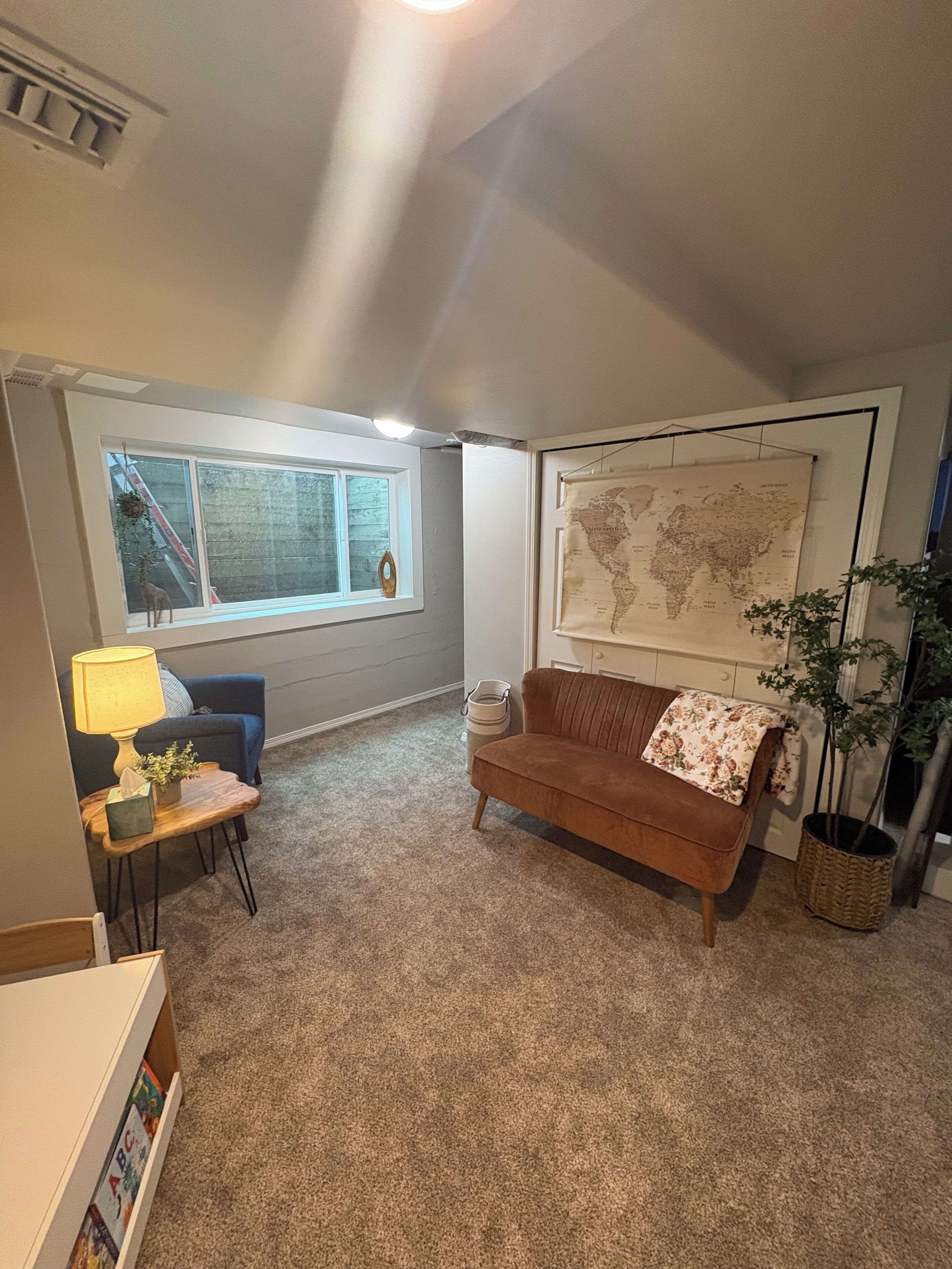 Un salon avec un canapé marron, une petite table avec une lampe, une carte du monde accrochée au mur et une fenêtre donnant sur un escalier.