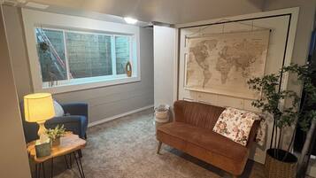 Un salon avec un canapé marron, une petite table avec une lampe, une carte du monde accrochée au mur et une fenêtre donnant sur un escalier.