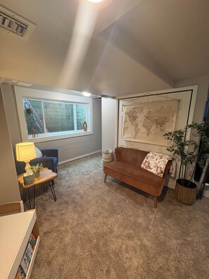 A living room with a brown sofa, a small table with a lamp, a world map on the wall, and a window with a view of a staircase.
