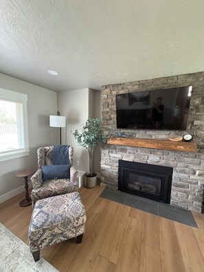 A living room with a stone fireplace, a television mounted on the wall, a patterned armchair, and a small table. - Spacious 4-bedroom, 5 bed, Family House with AC in perfect Missoula location (Missoula)