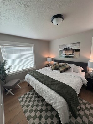 A bedroom with a bed, bedside lamps, a window with blinds, a painting on the wall, and a ceiling light.