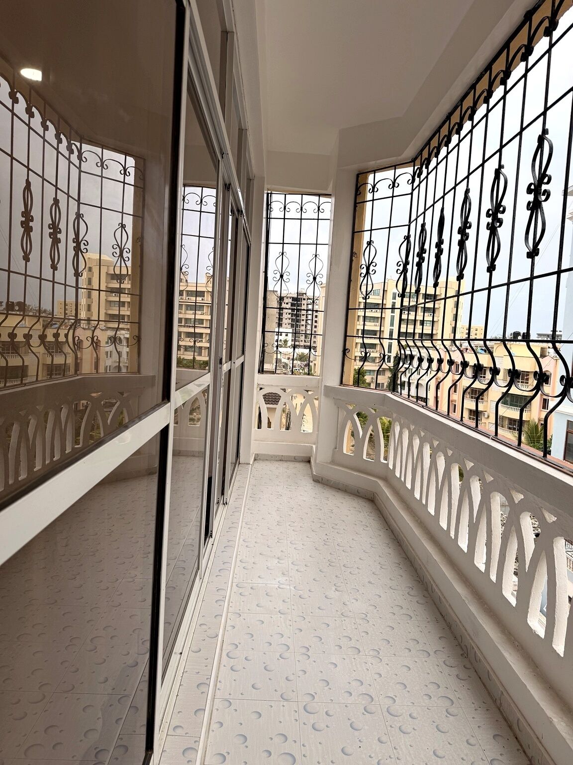 A balcony with patterned floor tiles, metal railings with decorative elements, and a view of surrounding buildings.