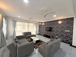 A modern living room with a sofa set, a coffee table, a TV mounted on the wall, and a ceiling fan. - Modern 3BR Nyali Apartment with AC, Wi-Fi and Backup Generator (Mombasa)