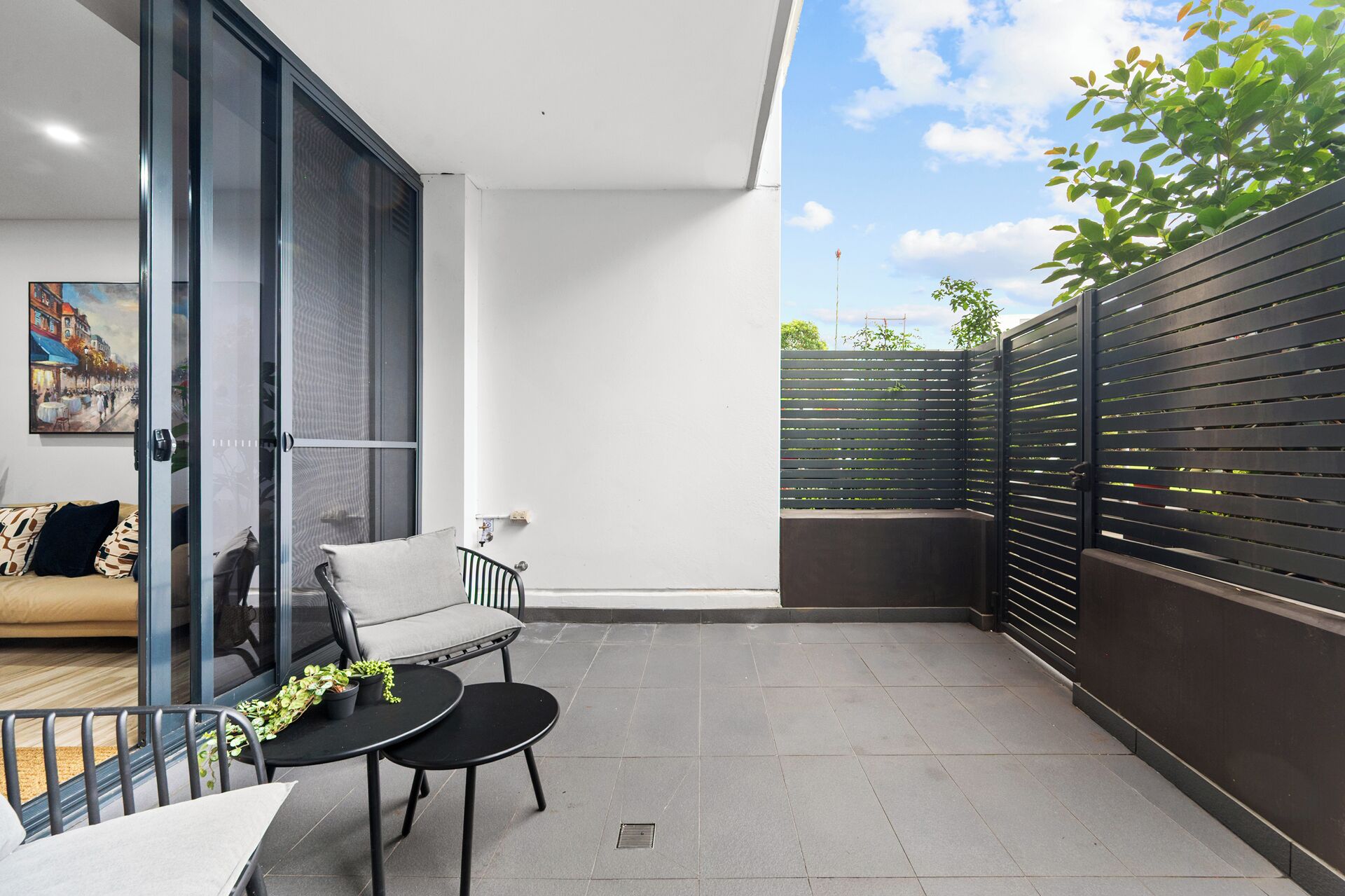 A modern balcony with a sofa, a small table with plants, and a view of the sky and greenery.