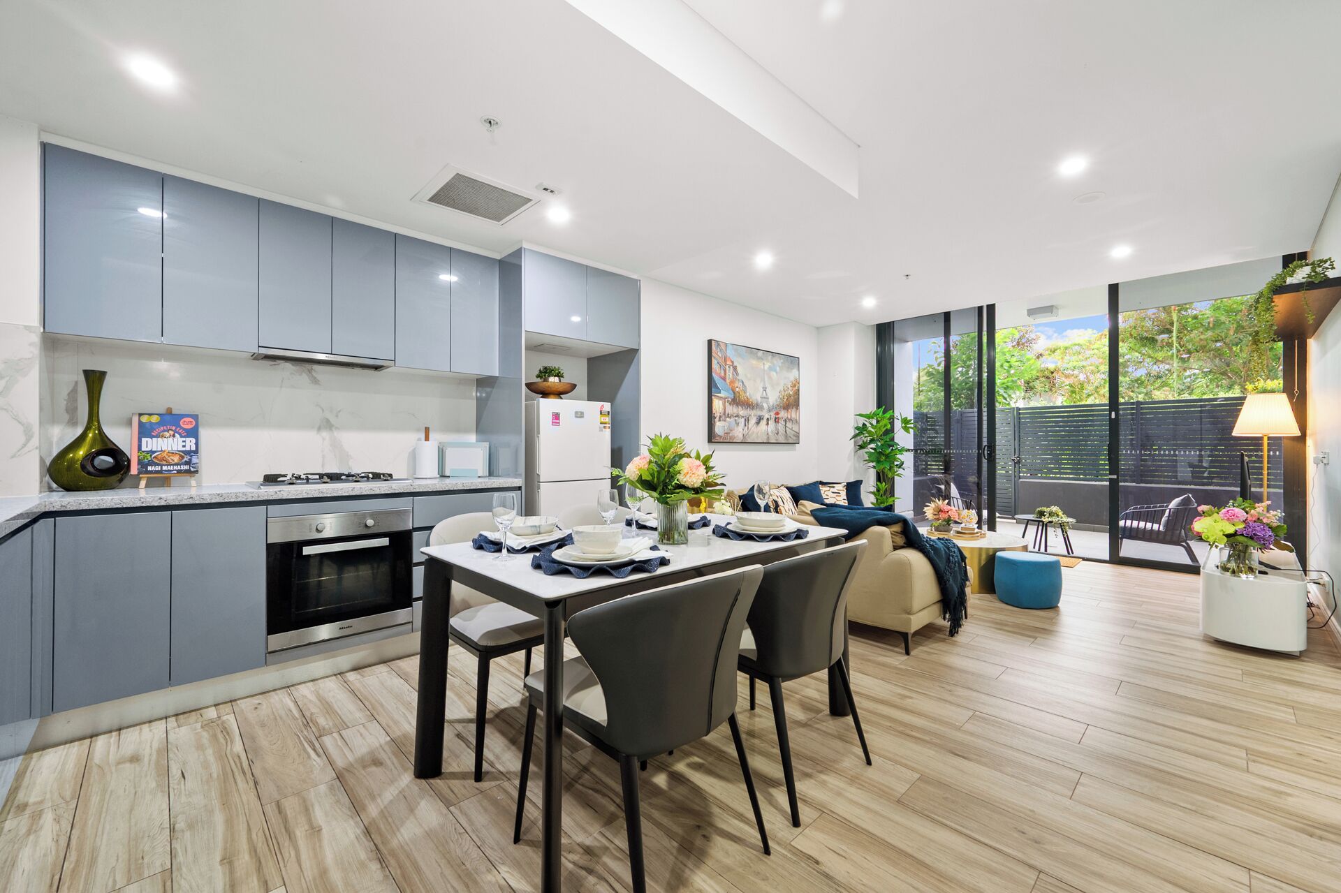 A modern kitchen with grey cabinets, a dining area with a table set for two, and a view of a garden through large sliding doors.