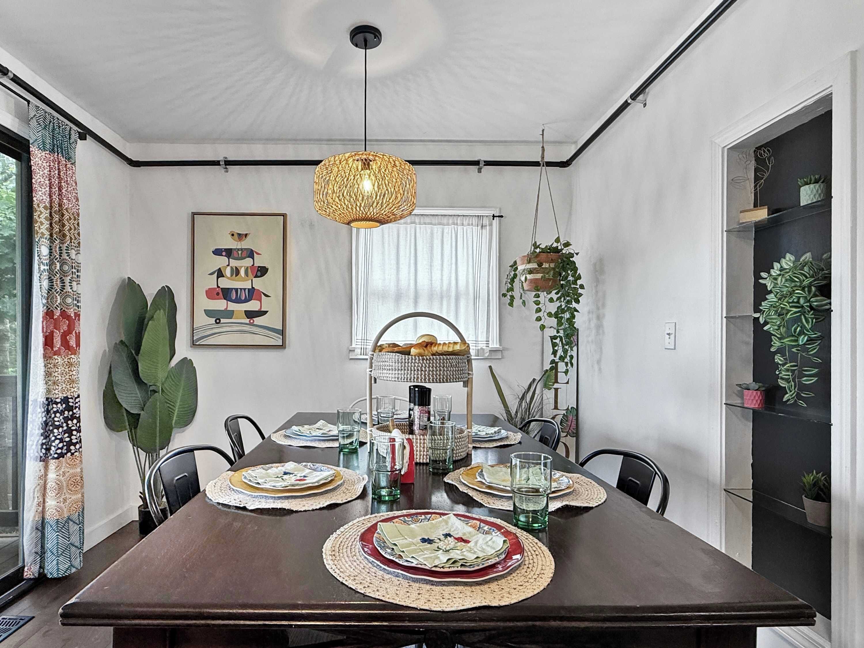 Un coin repas avec une table en bois foncé, dressée pour quatre personnes et entourée de chaises. Un luminaire suspendu se trouve au-dessus de la table, et une fenêtre avec des rideaux est visible sur le côté gauche.