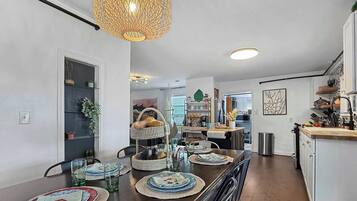 A dining area with a wooden table set for four, a hanging light fixture, and a kitchen in the background.