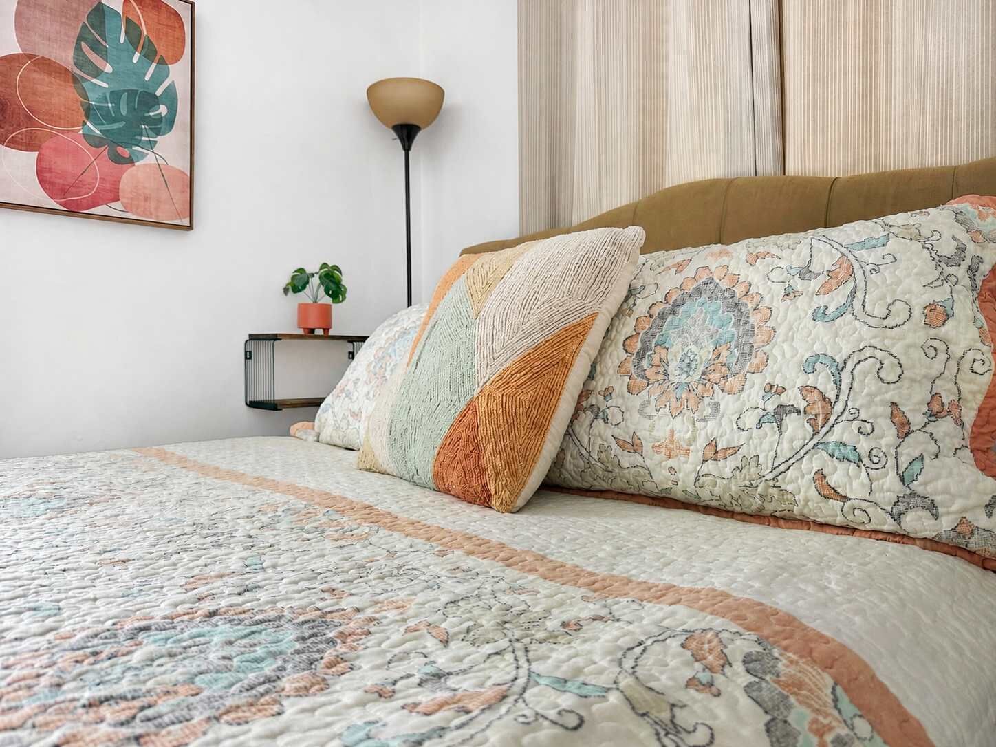 Une chambre à coucher avec un lit, des oreillers, une table de chevet ornée d’une plante et une lampe fixée au mur.