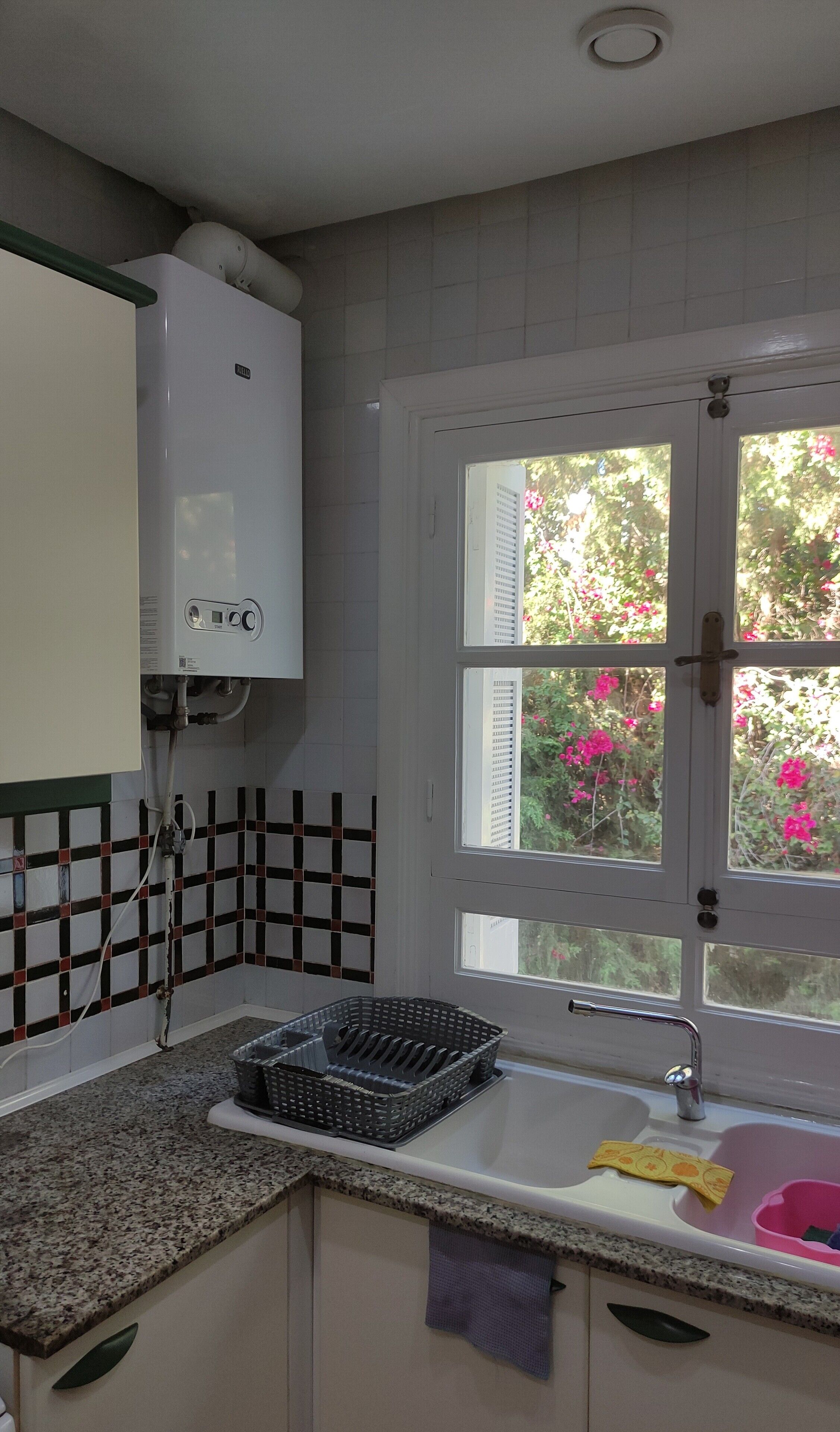 A kitchen with a water heater, a window with a view of pink flowers, a sink, and a granite countertop.