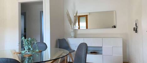 Una sala moderna con una mesa de comedor de vidrio, dos sillas grises, un armario blanco y un espejo grande.