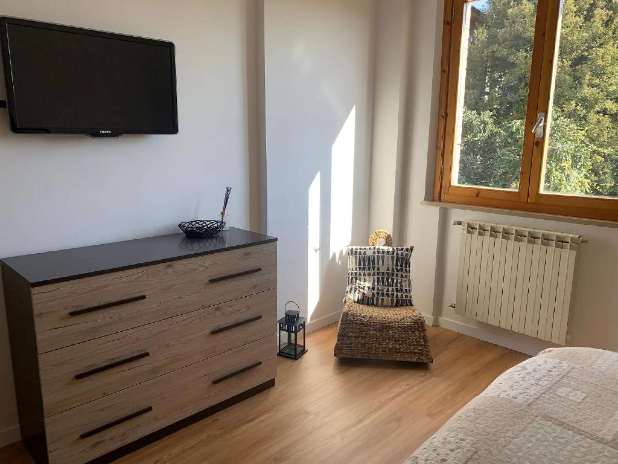 Un dormitorio con un aparador de madera, una mesita de noche con lámpara, un radiador y una ventana con vistas a árboles.