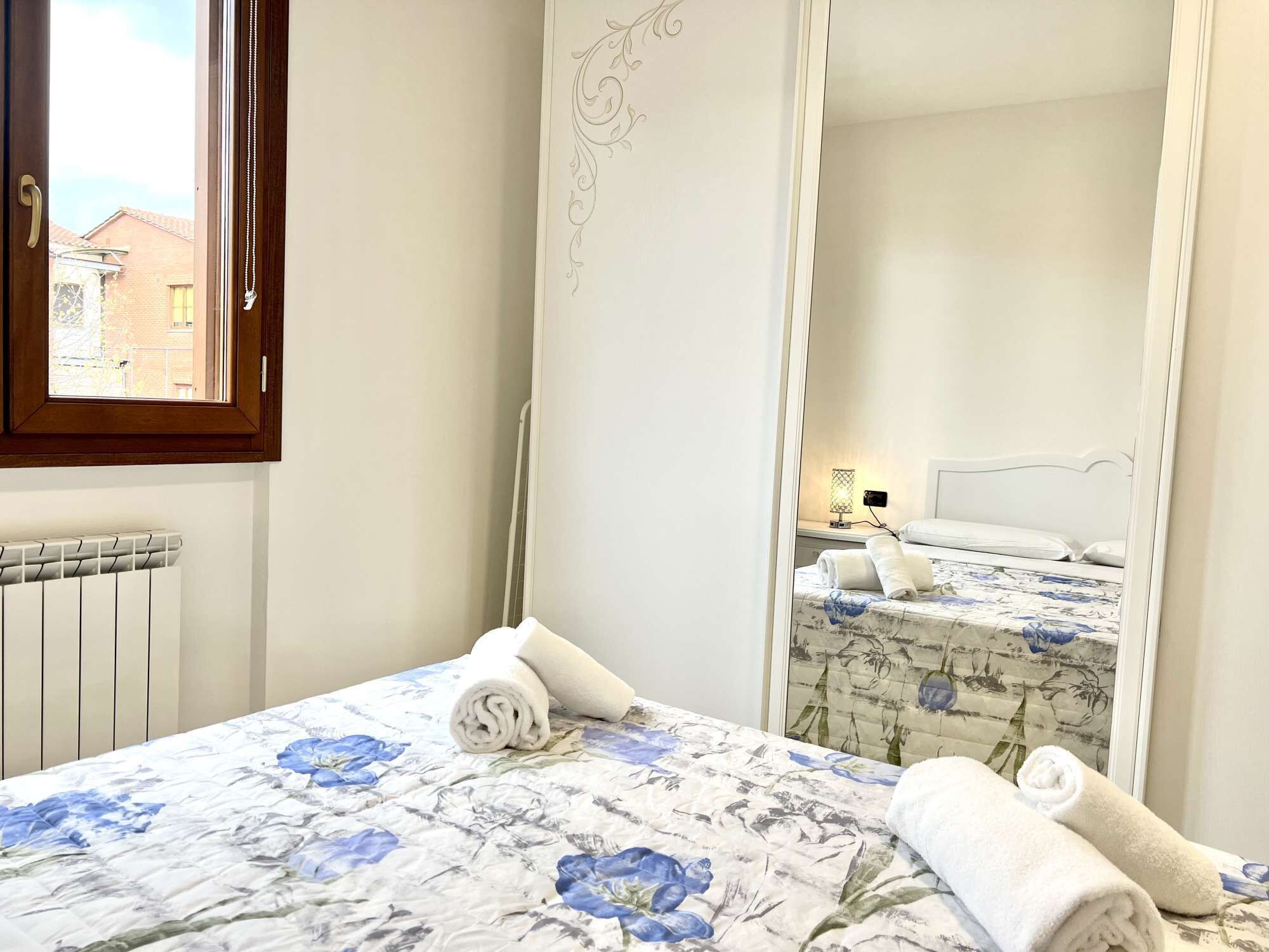 A hotel room with a bed, a mirror, a radiator, and a window with a view of buildings.