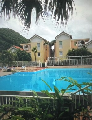 Ein Swimmingpool mit weißem Zaun, umgeben von Grünpflanzen und Gebäuden mit vielen Fenstern.